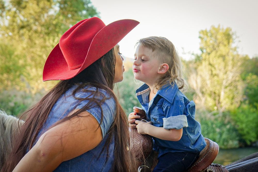 A woman in a red cowboy hat shares a sweet moment with a young child in a denim shirt outdoors.
