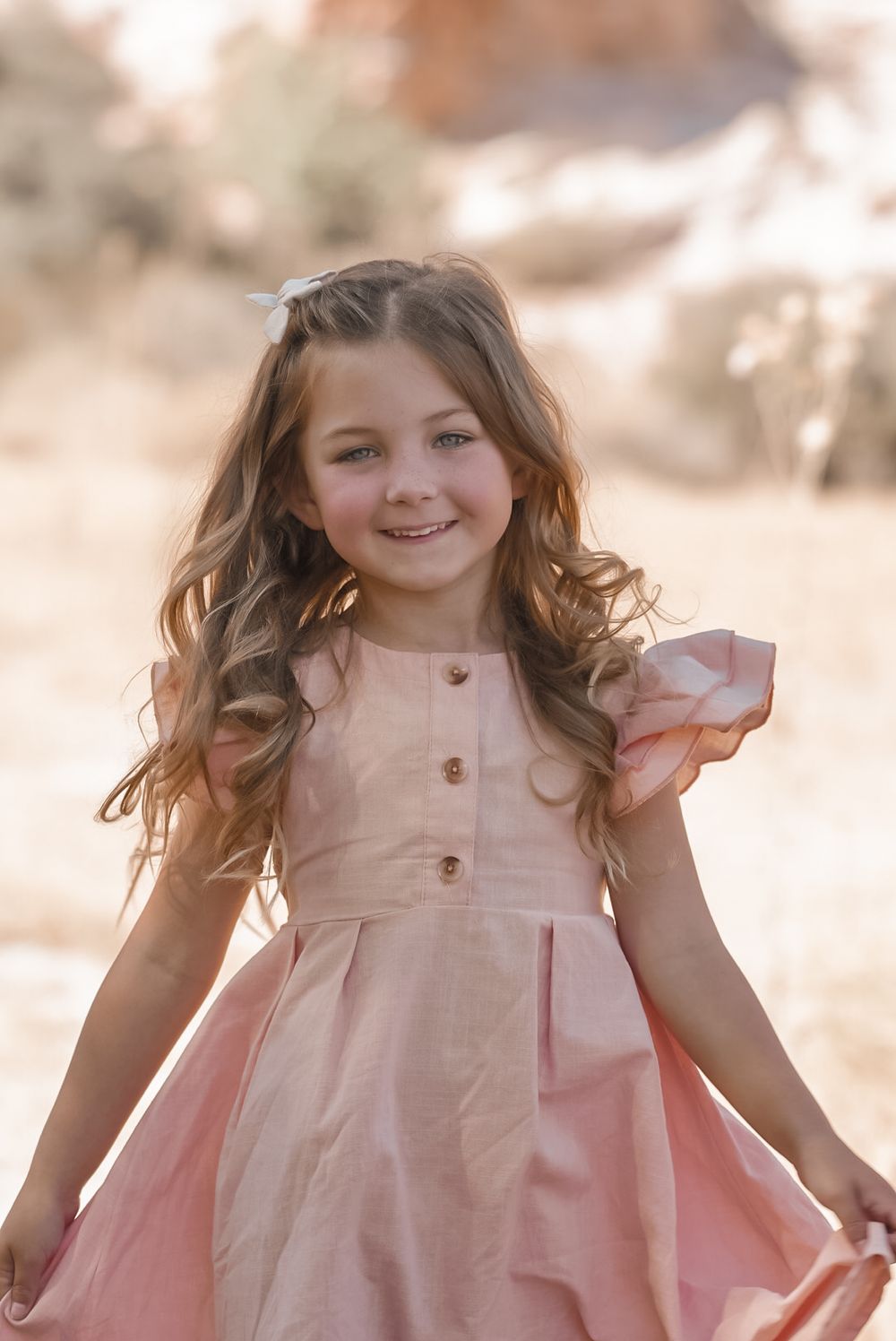 Adorable young girl in a pink dress smiling outdoors in a sun-drenched natural setting.