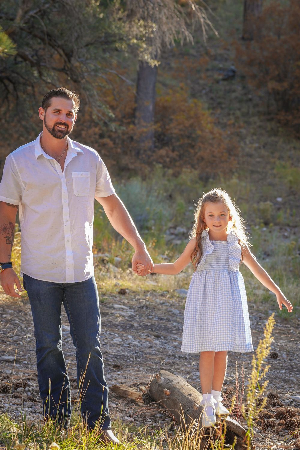 A bearded father in a white shirt holds hands with his young daughter in a blue dress outdoors in a sunlit forest.