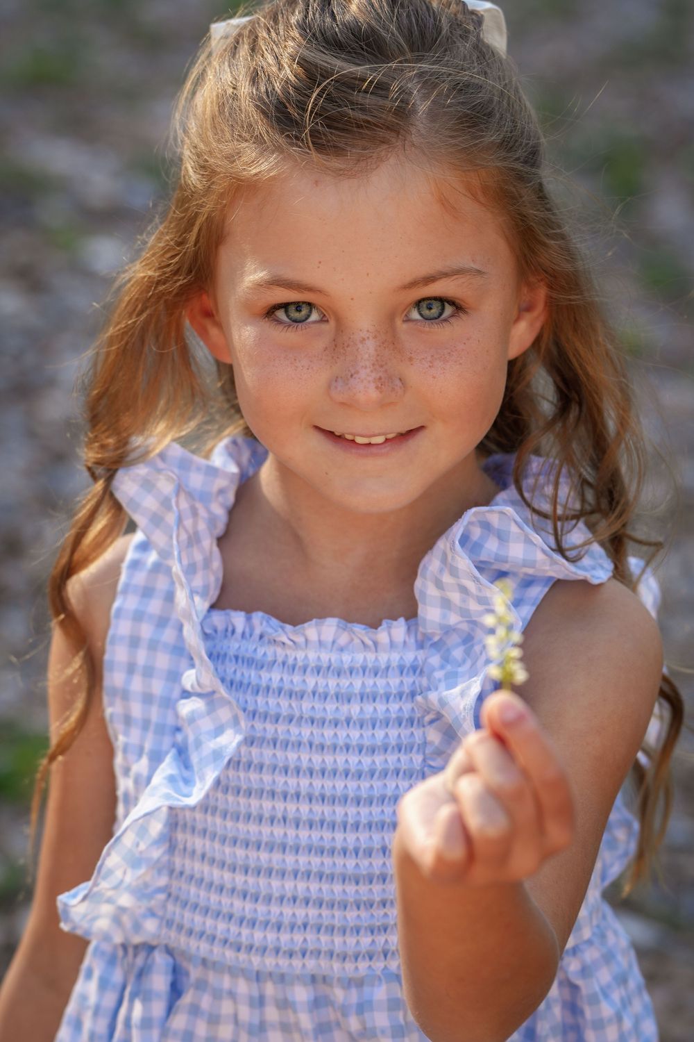 Young girl with freckles in blue gingham dress holds a small flower, smiling outdoors in warm sunlight.