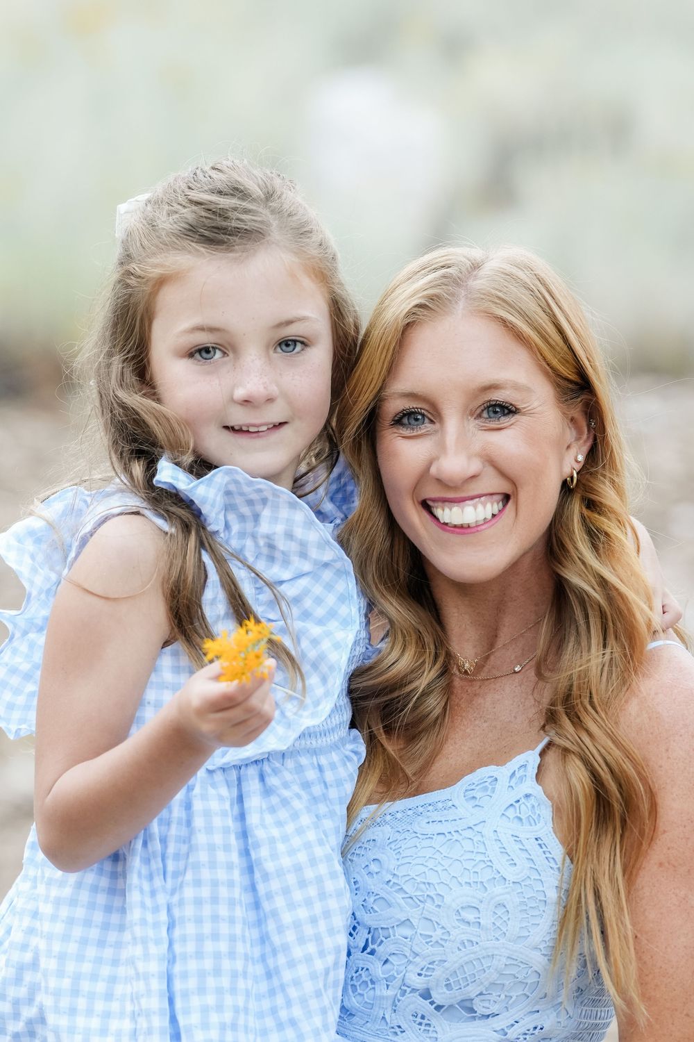 A smiling woman and young girl in matching blue outfits pose together outdoors, the child holding a yellow flower.