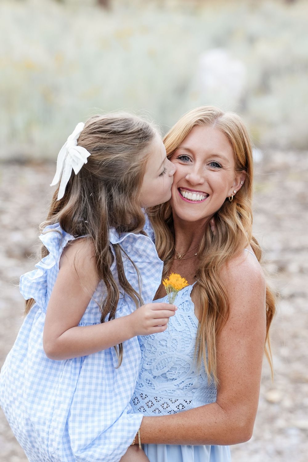 A young girl in a blue gingham dress kisses a smiling blonde woman holding an orange flower outdoors.