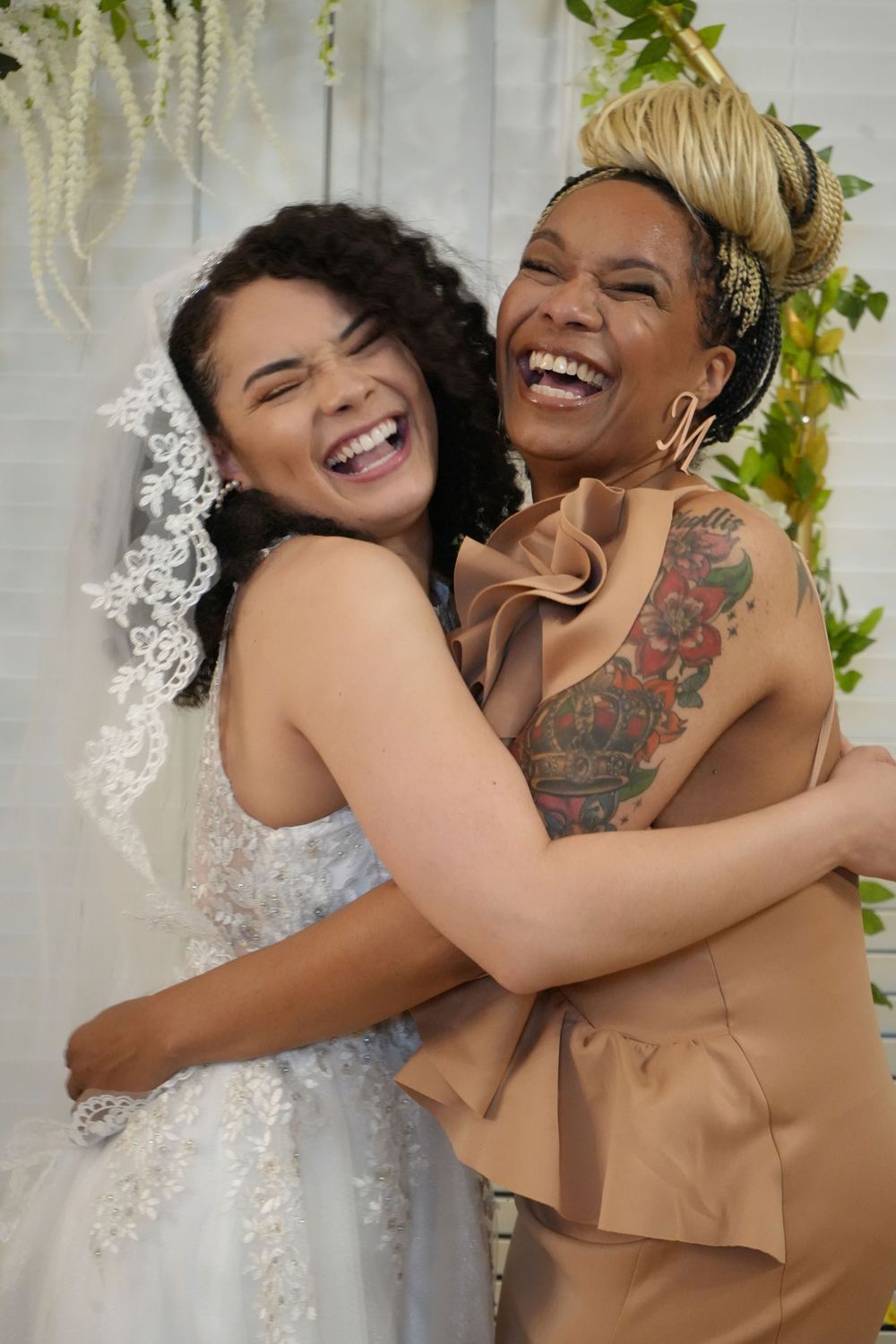 Two joyful women hugging and laughing at a wedding, one in a white bridal gown with veil, the other in a tan outfit with tattoos.