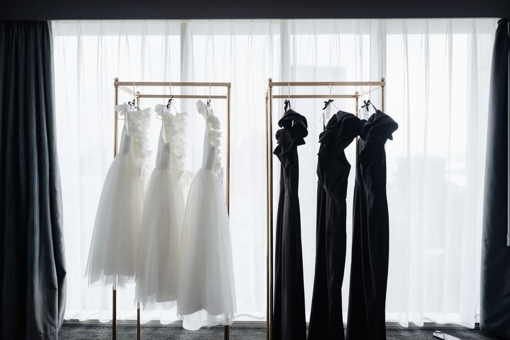 White wedding gowns and black bridesmaid dresses hanging on gold clothing racks near a bright window.