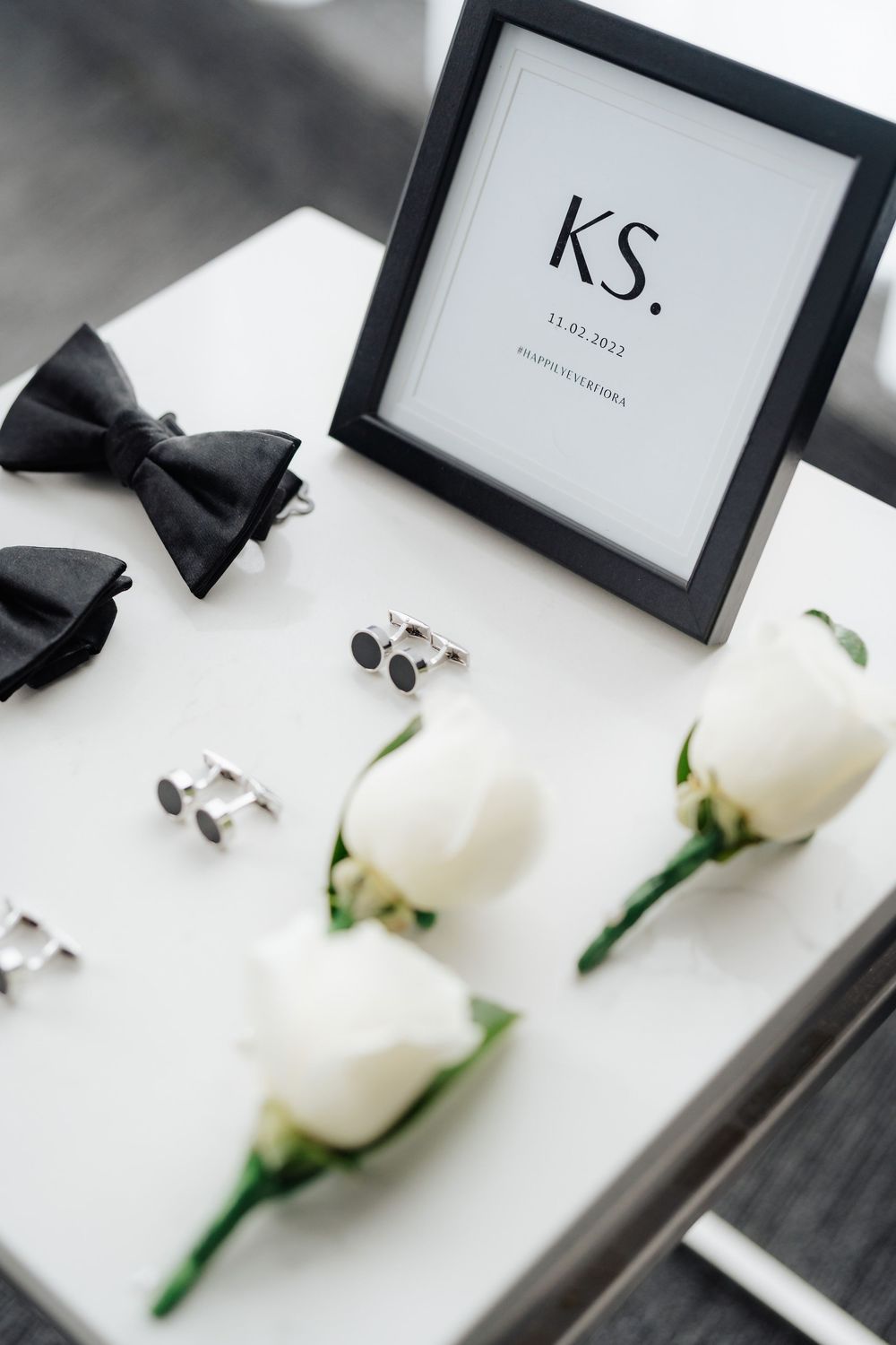 Elegant wedding flat lay with white roses, black bow tie, cufflinks, and framed KS monogram on white surface.