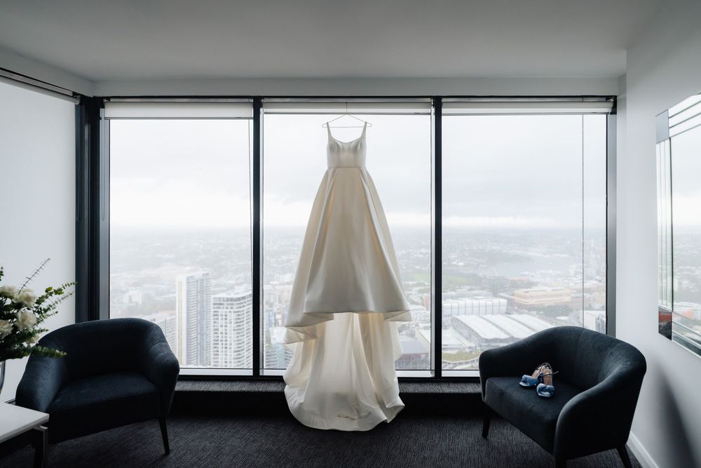A elegant white wedding dress hanging in front of floor-to-ceiling windows with a city skyline view.
