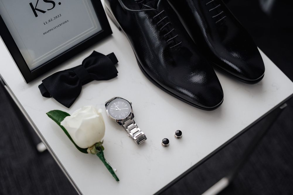 Elegant groom accessories flat lay featuring black Oxford shoes, bow tie, silver watch, cufflinks, and white rose on white surface.