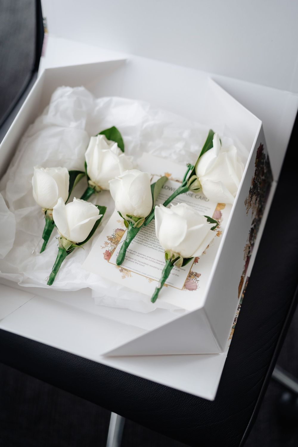 White rose boutonnieres arranged in an open gift box with tissue paper, perfect for a wedding or formal event.