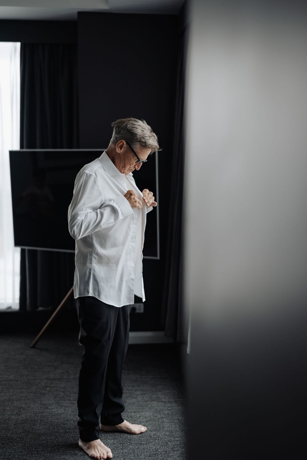 Elderly man in white shirt and black pants buttoning up indoors, standing barefoot on gray carpet.