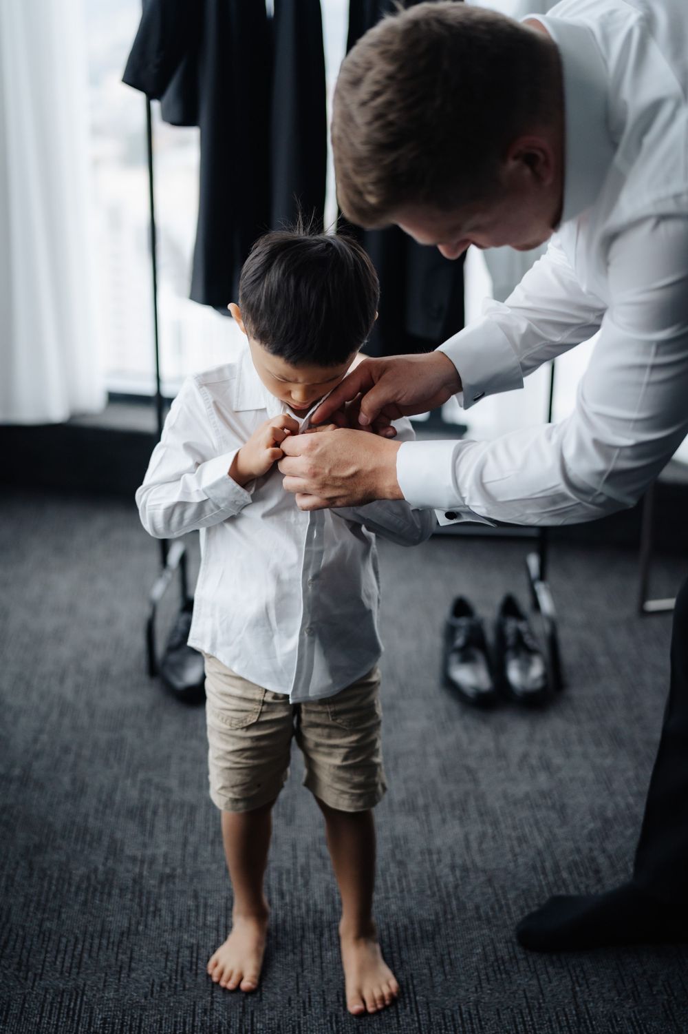 A father helps his young barefoot son button up a white dress shirt in a modern room with shoes on the floor.
