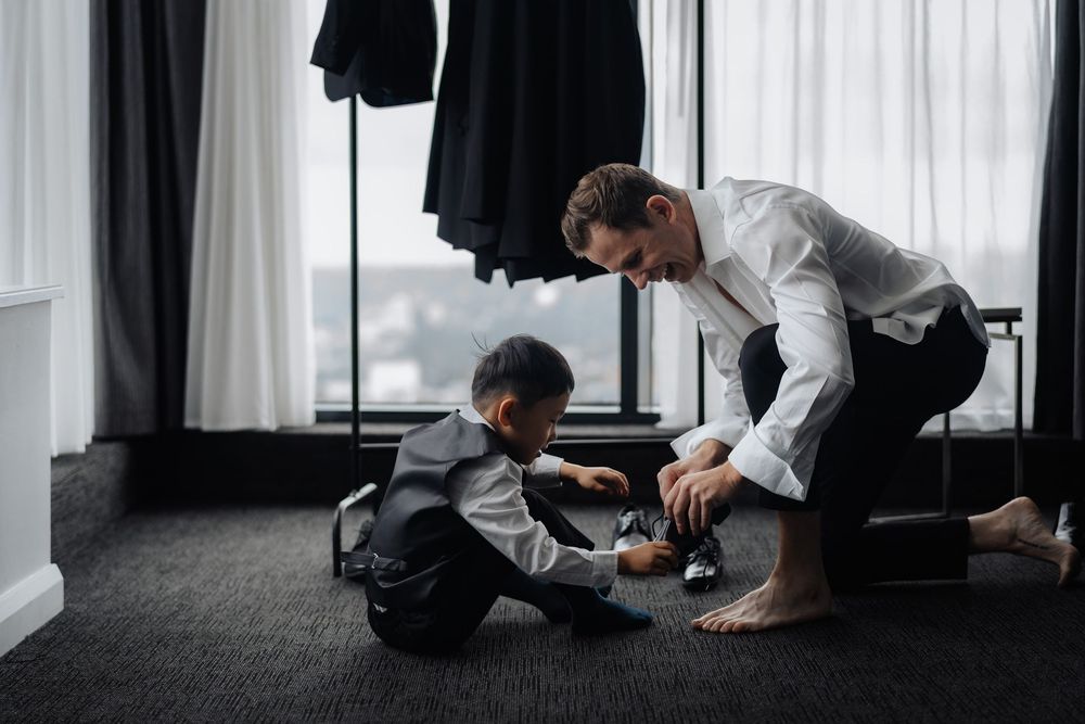 A man in a white shirt helps a young boy in a suit put on shoes in a hotel room before a wedding.