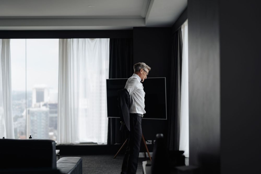 A businessman in a white shirt stands by a large window in a modern hotel room with dark decor and city views.