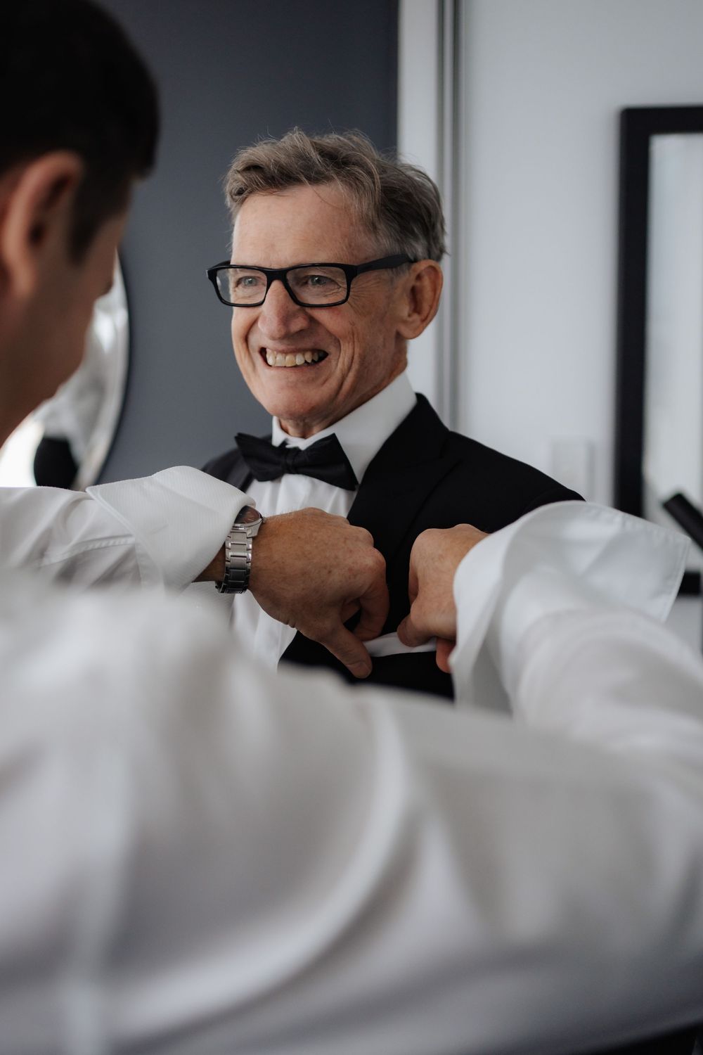 Smiling older man in tuxedo with bow tie and glasses receiving help adjusting cufflinks from another man in formal wear.