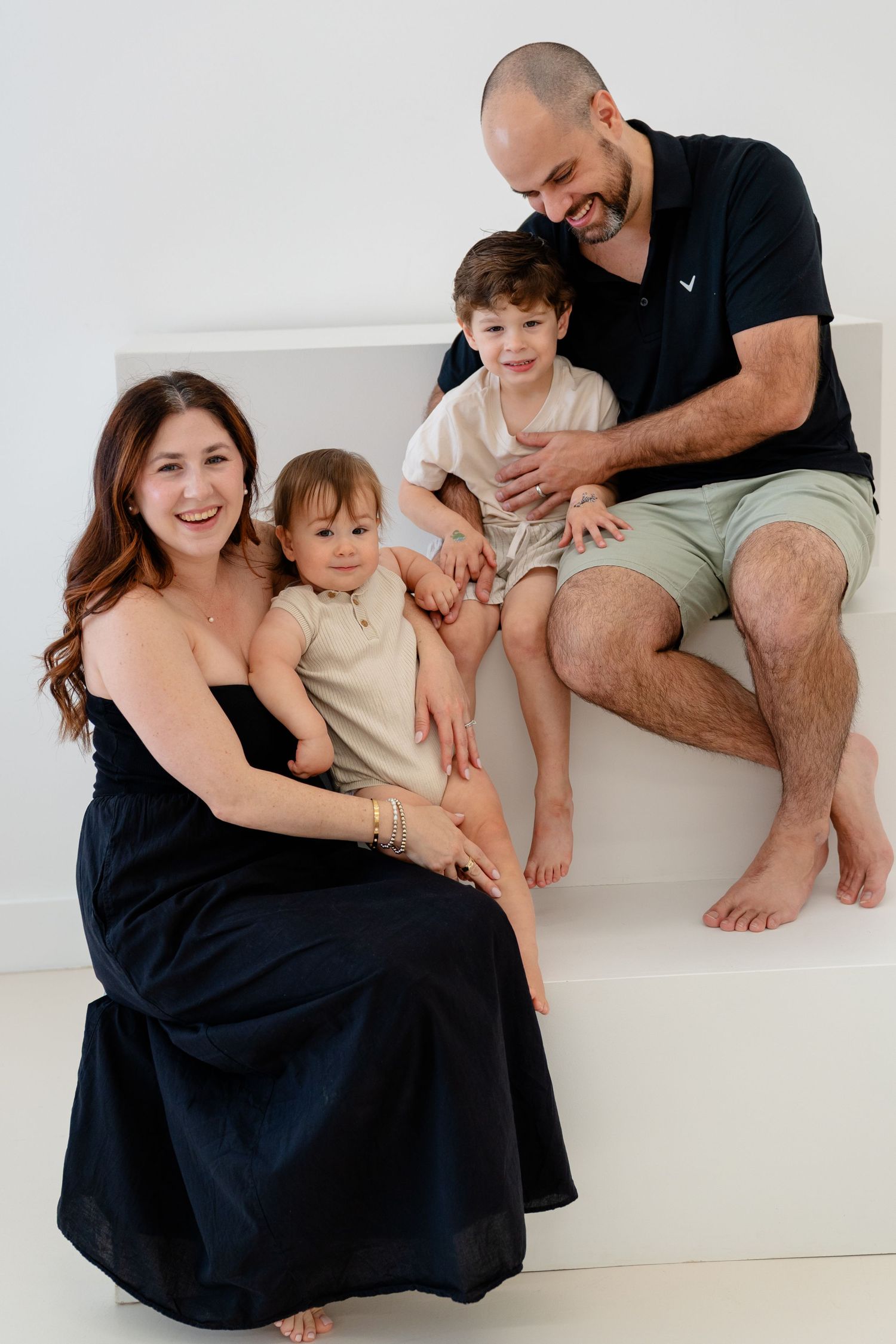 A happy family of four poses together on white steps, with mom in a black dress and dad in a navy polo with two young children.