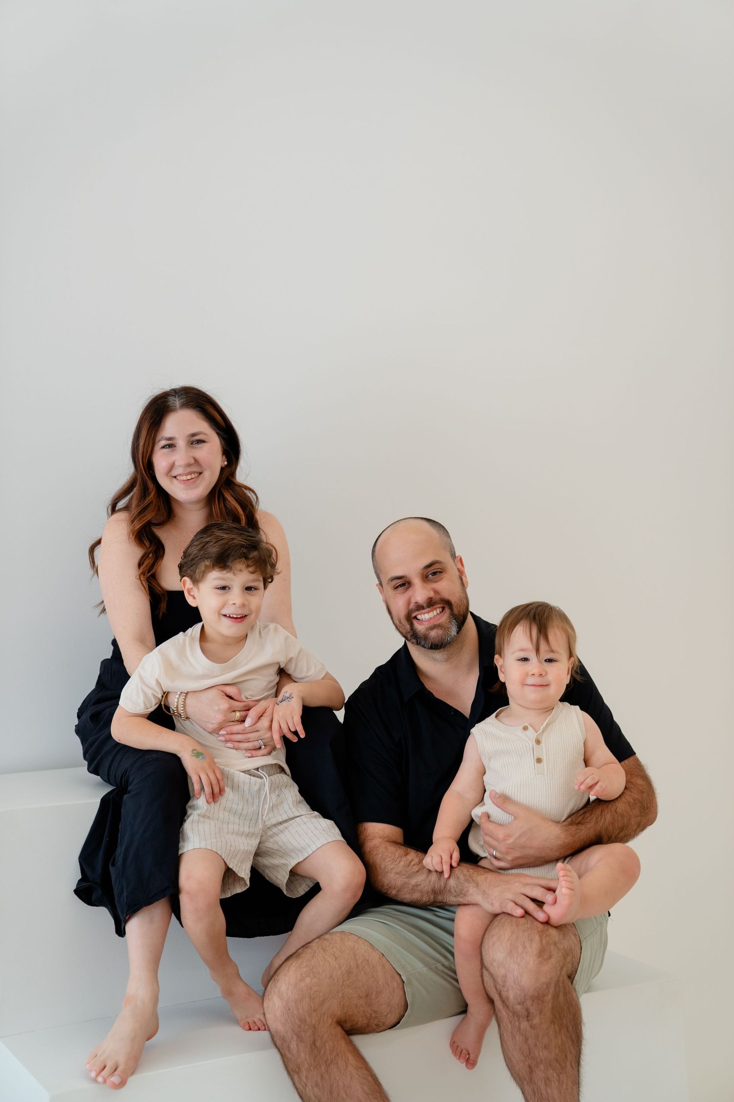 Family of four posing together, parents smiling with two young boys in a bright white studio setting.