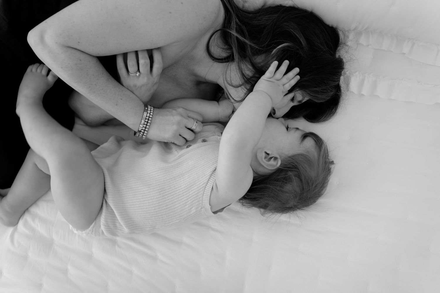 Black and white photo of a mother tenderly kissing her baby while lying on a white bed.