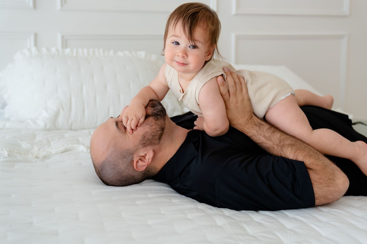 A loving father lies on a white bed playfully holding his smiling baby in a cozy indoor setting.