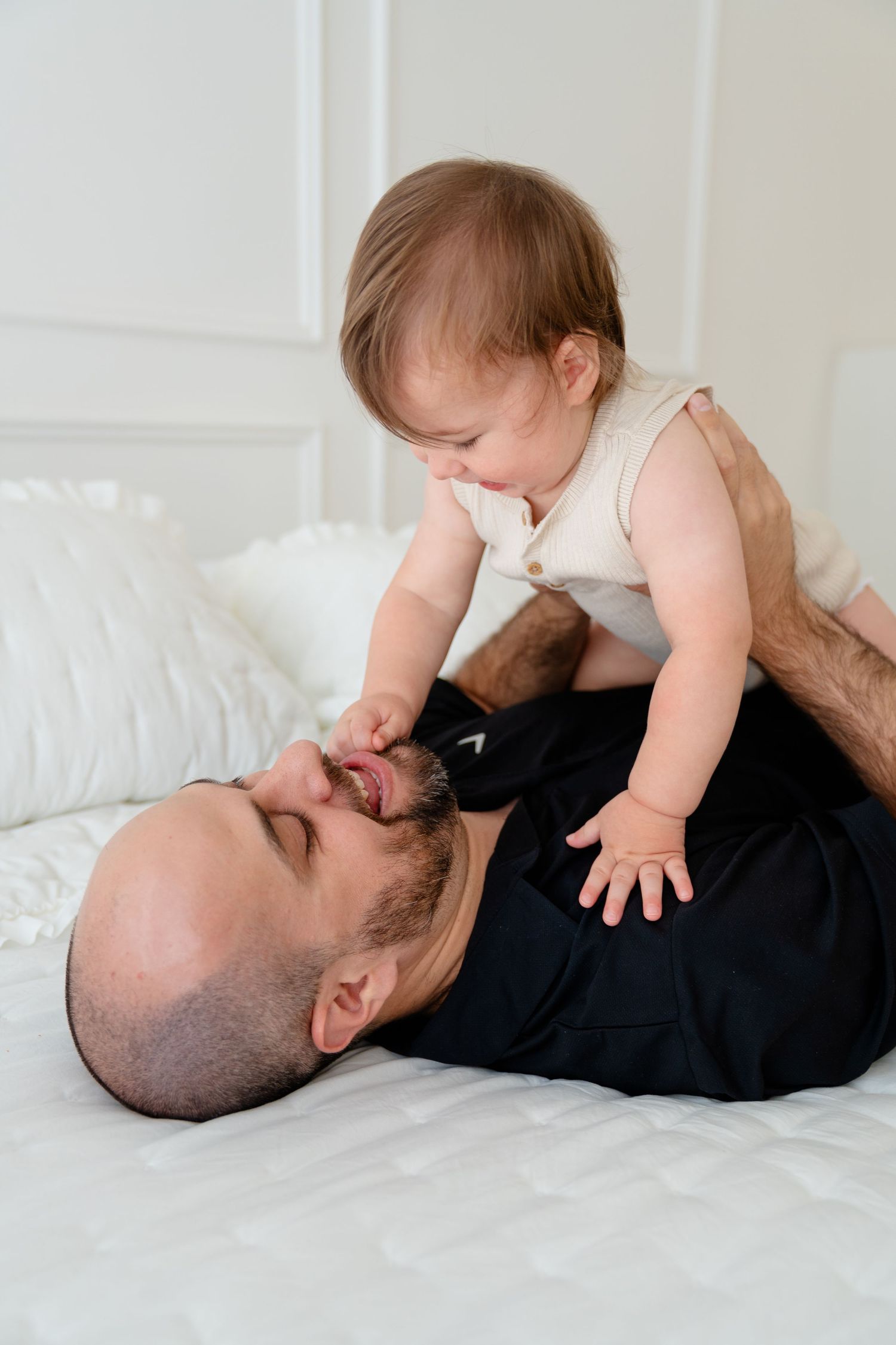 Father lying on bed playfully kissing his smiling baby while holding the infant up above his chest.