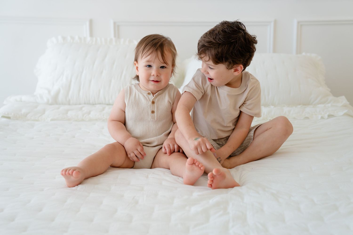 Two young siblings in matching beige outfits sitting together on a white bed, smiling and playing.