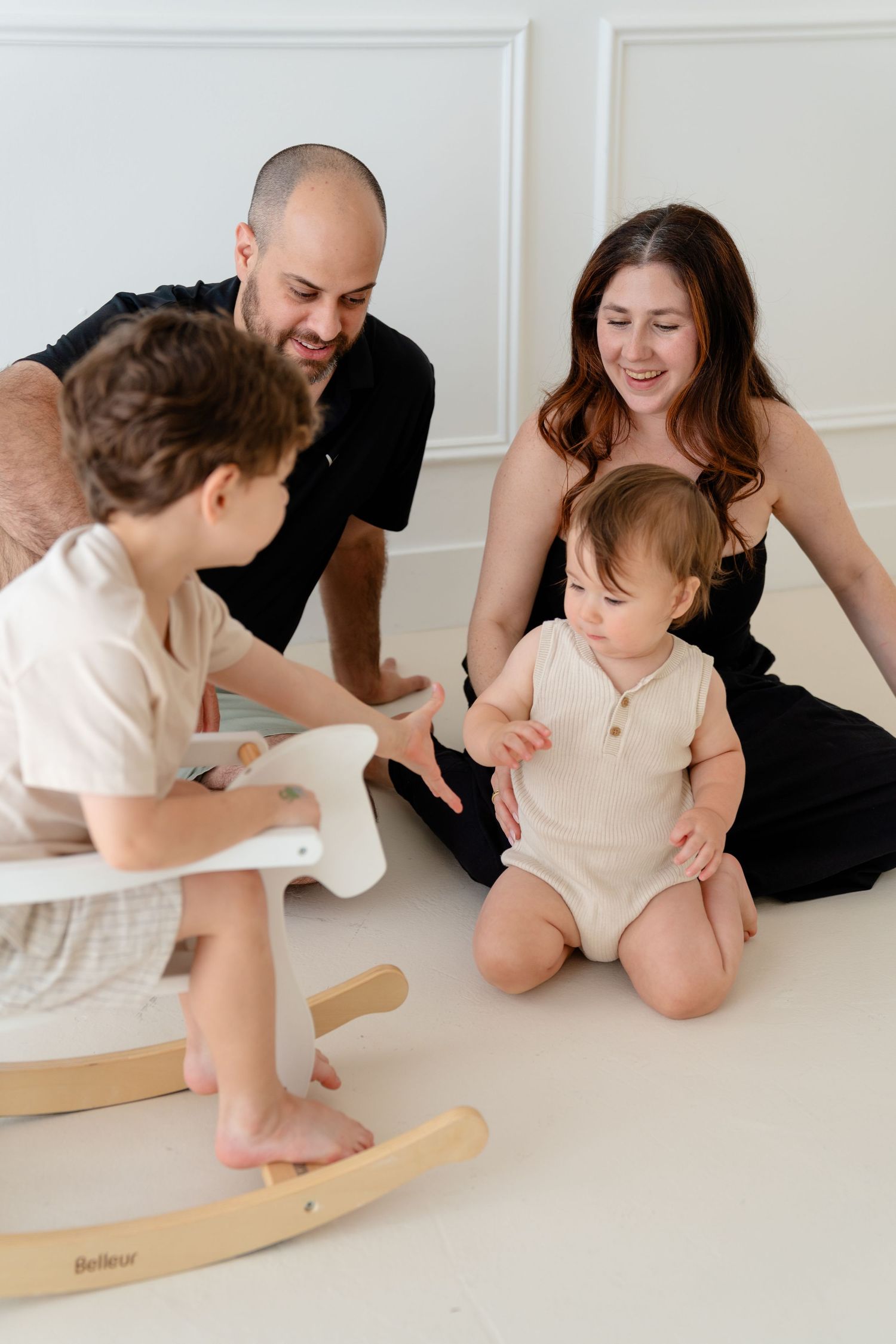 Family of four with two young children playing together on white floor, parents smiling at baby in beige onesie.