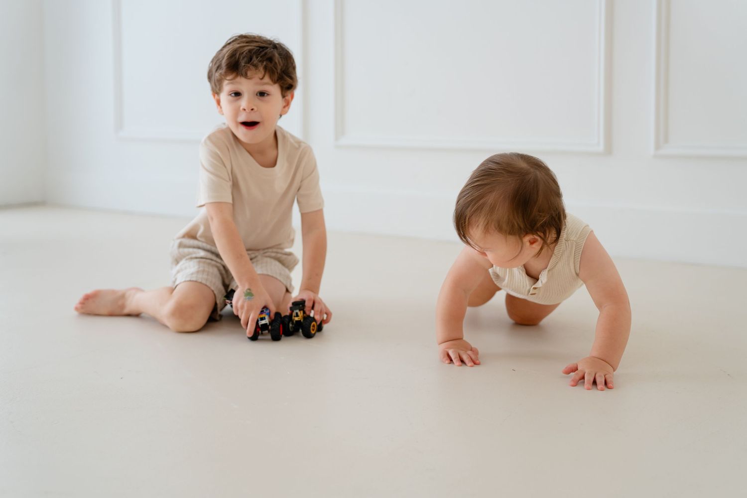 Two young children playing on a white floor, one sitting with toy cars and one crawling.