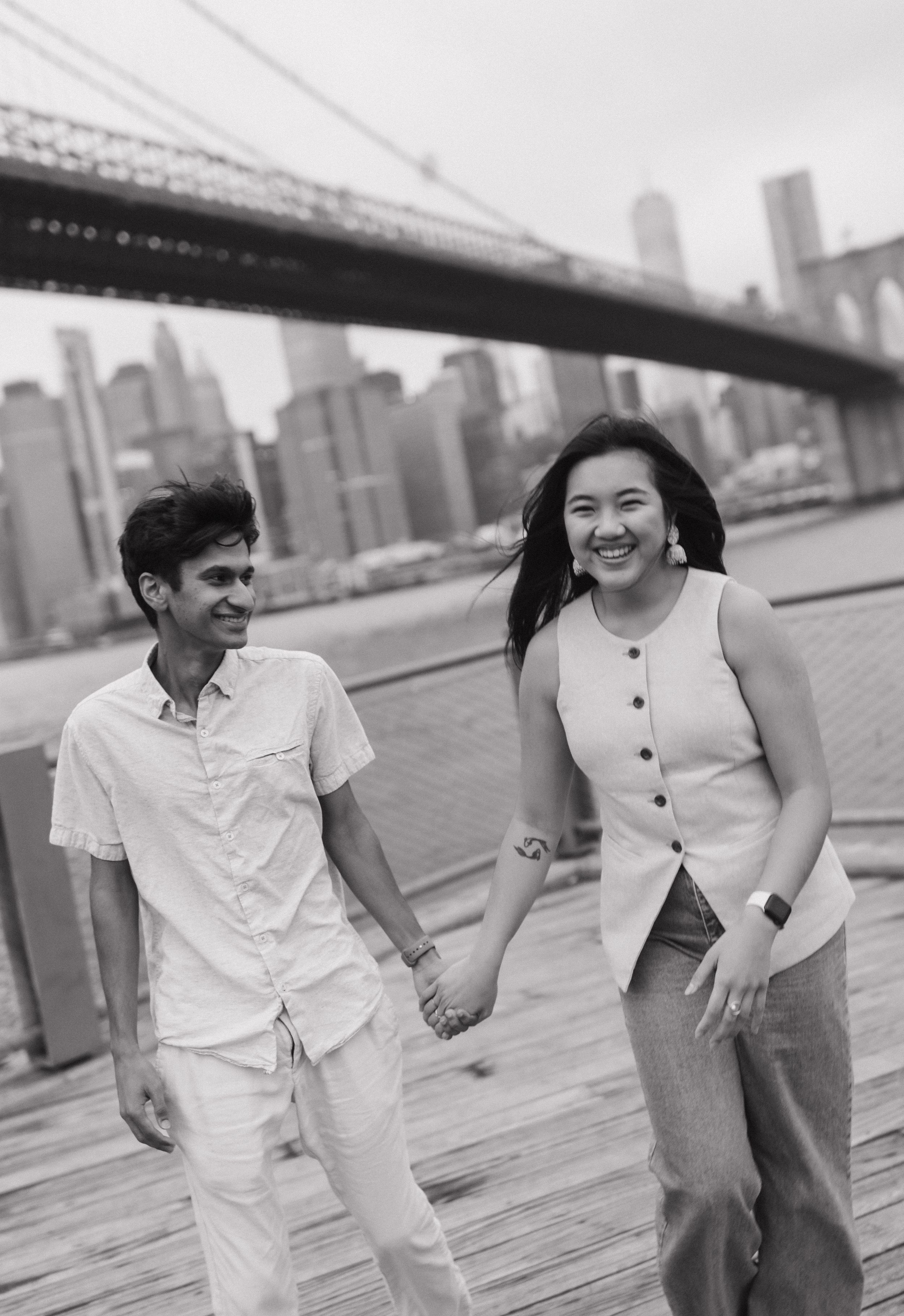 A smiling couple holds hands on a wooden boardwalk with the Brooklyn Bridge and NYC skyline in the background.