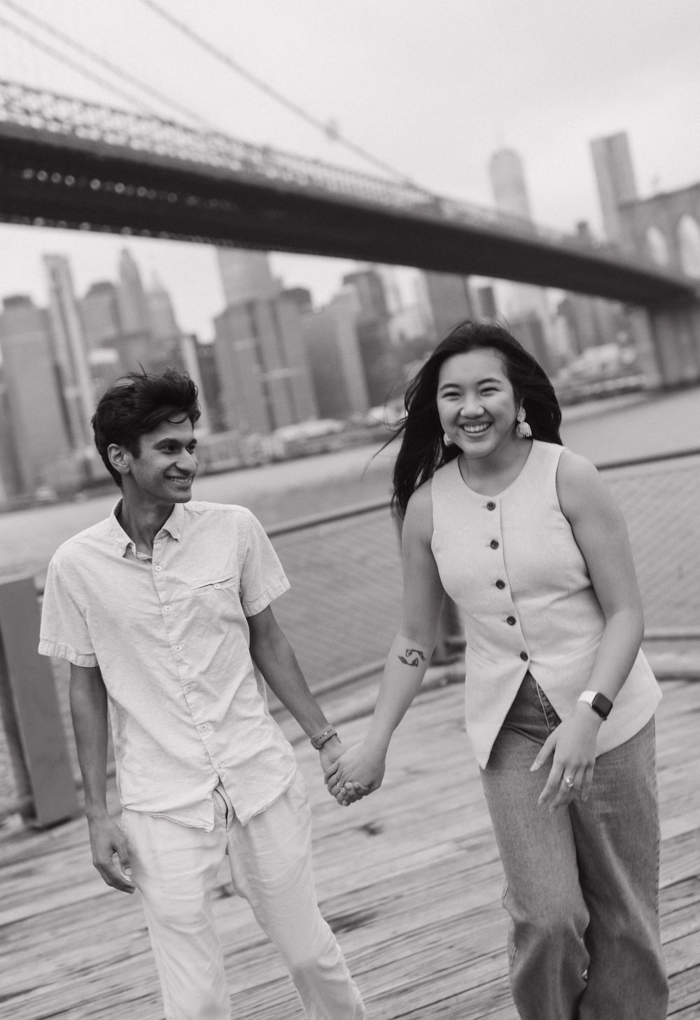 A smiling couple holds hands on a wooden boardwalk with the Brooklyn Bridge and NYC skyline in the background.
