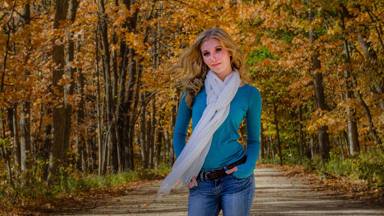 A young blonde woman in a teal sweater and white scarf poses on a forest path surrounded by vibrant autumn foliage.