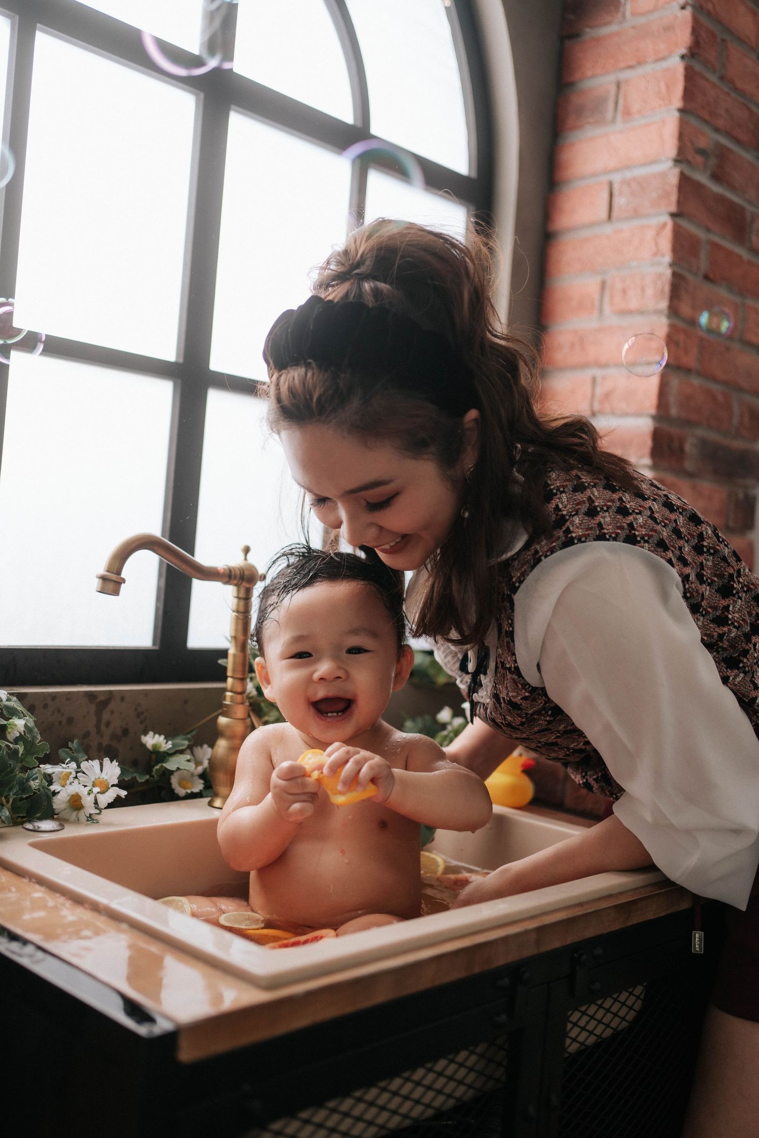 A smiling mother bathes her happy baby in a kitchen sink near a large window with brick walls.一位微笑的母親在廚房水槽旁,對著一扇磚牆大窗戶,為快樂的孩子洗澡。