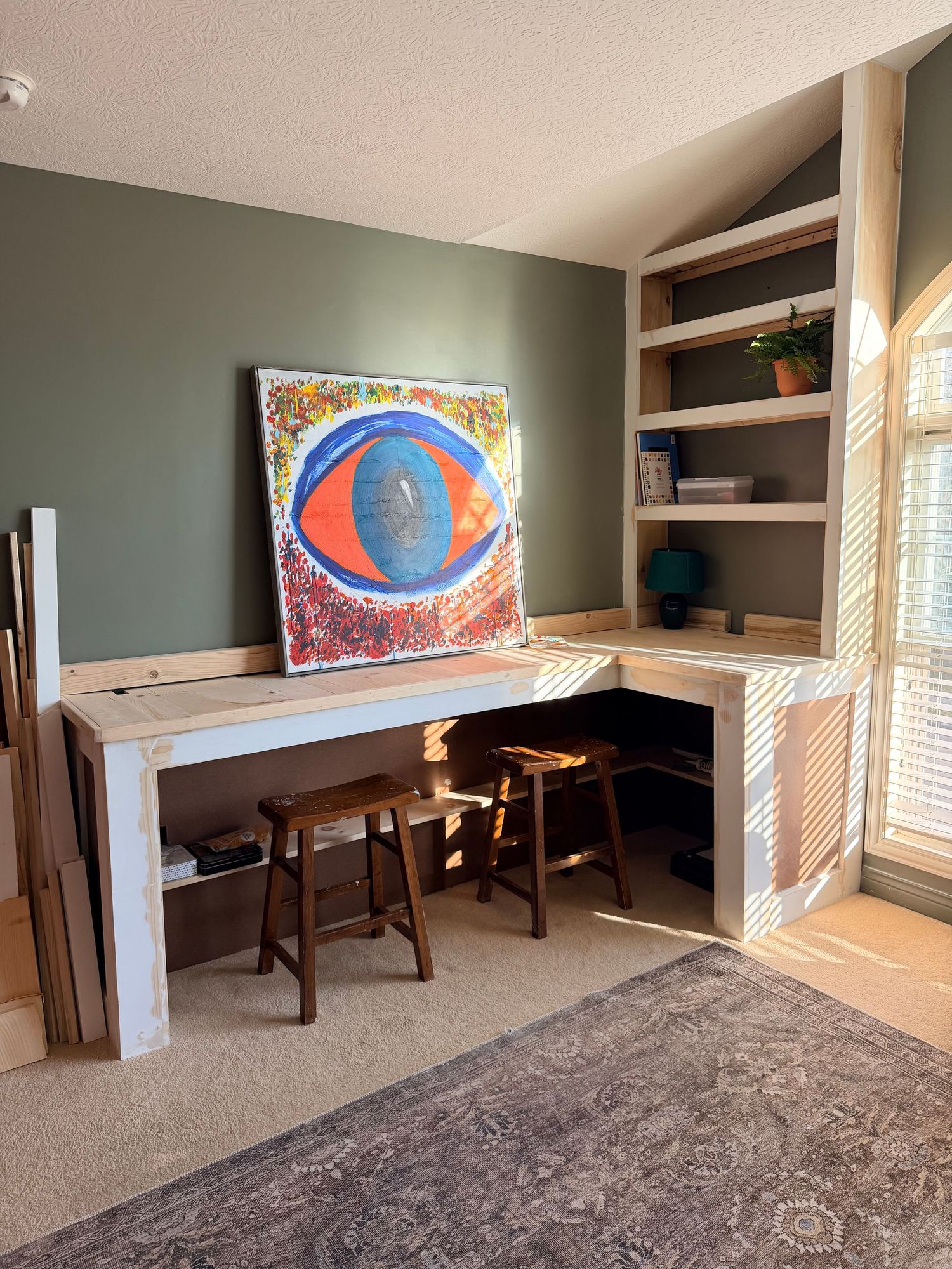 Custom built-in corner desk with open shelving, two wooden stools, and colorful eye artwork on sage green wall.