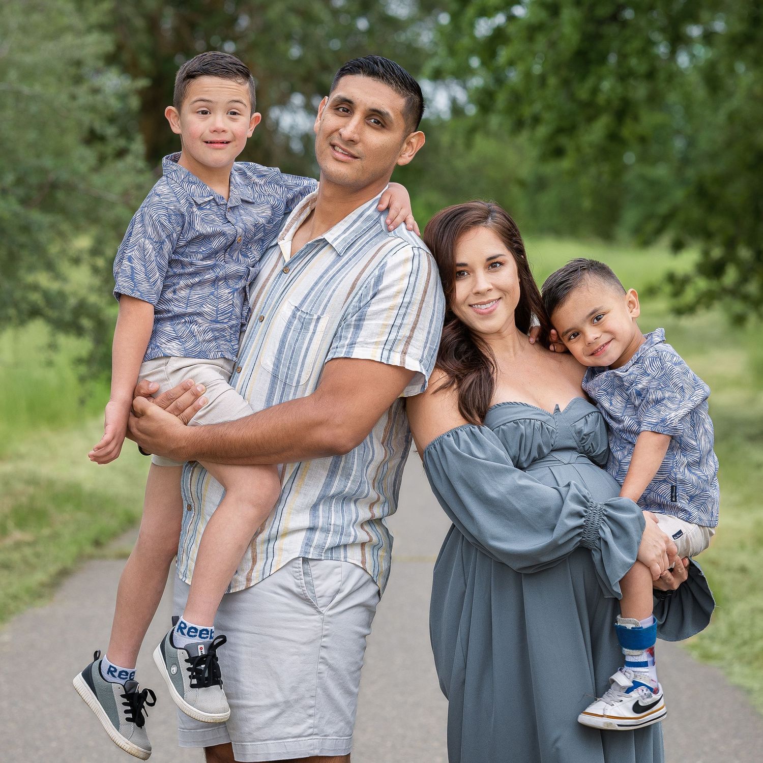 Happy family of four posing outdoors on a tree-lined path, parents holding two young boys in coordinating outfits.