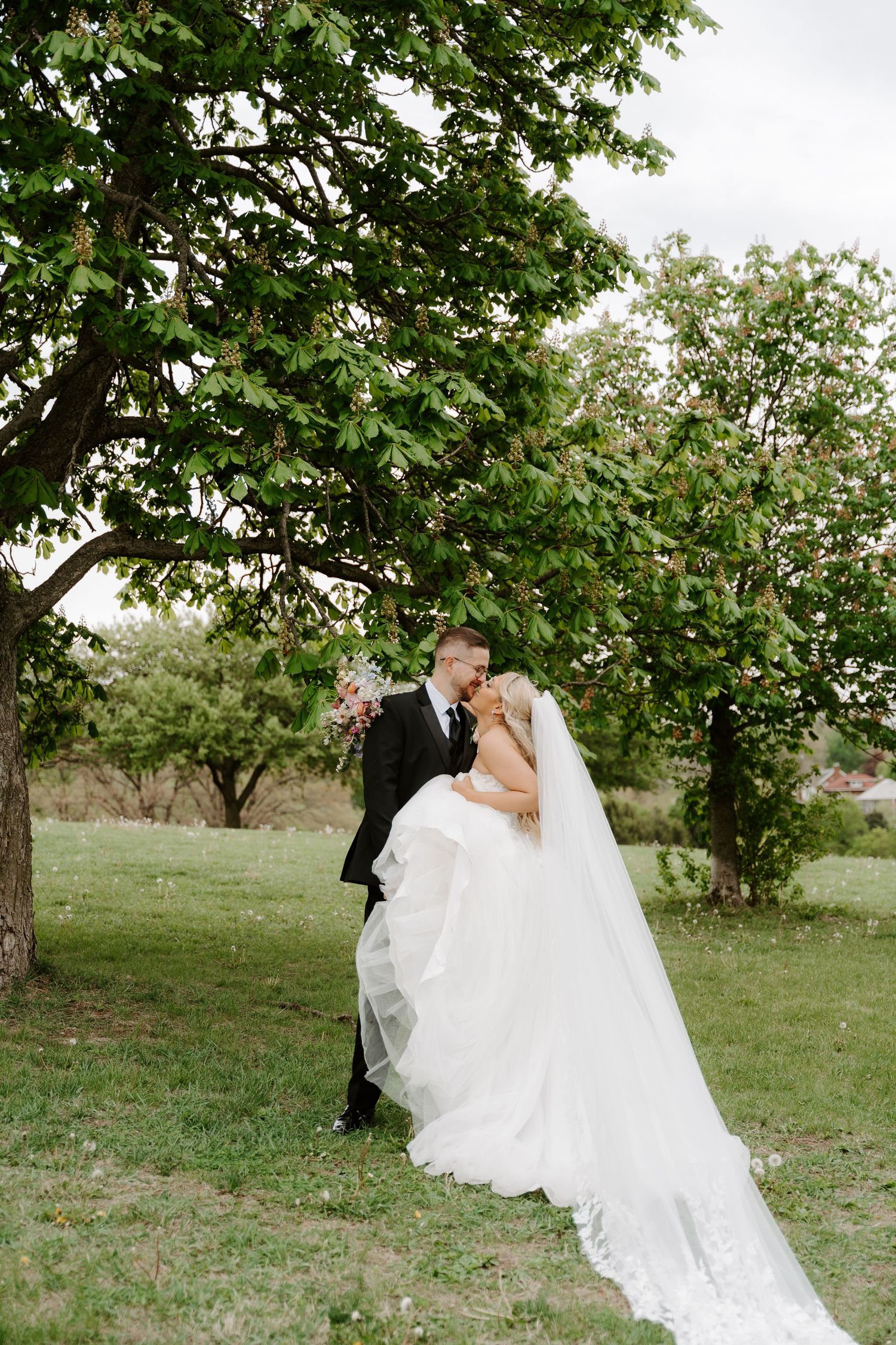 Black and white photo of a bride and groom sharing a romantic kiss under a large tree in a lush garden.
