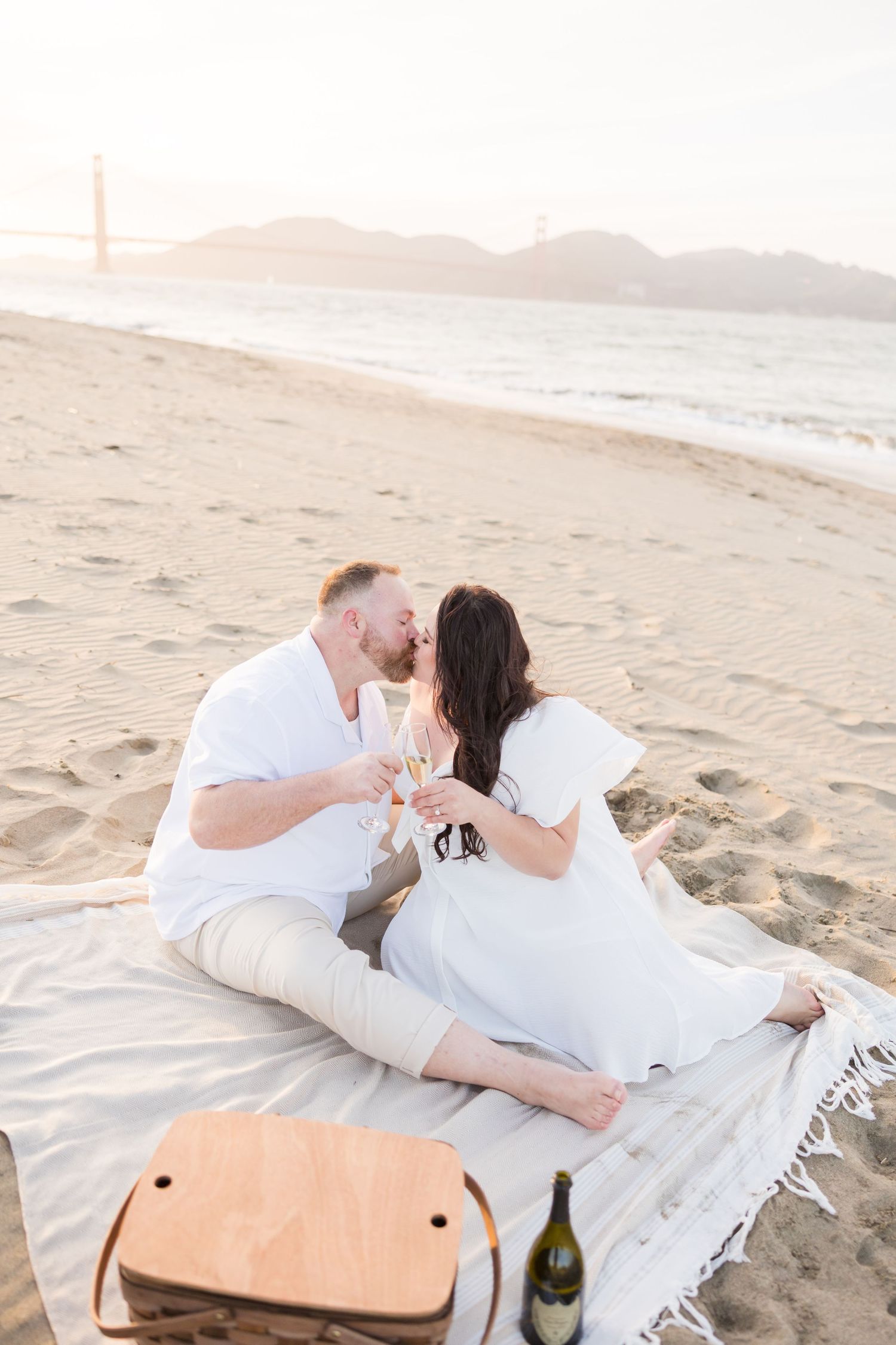 Couple kissing on Baker Beach with Golden Gate Bridge backdrop, white outfits, picnic basket, champagne bottle.