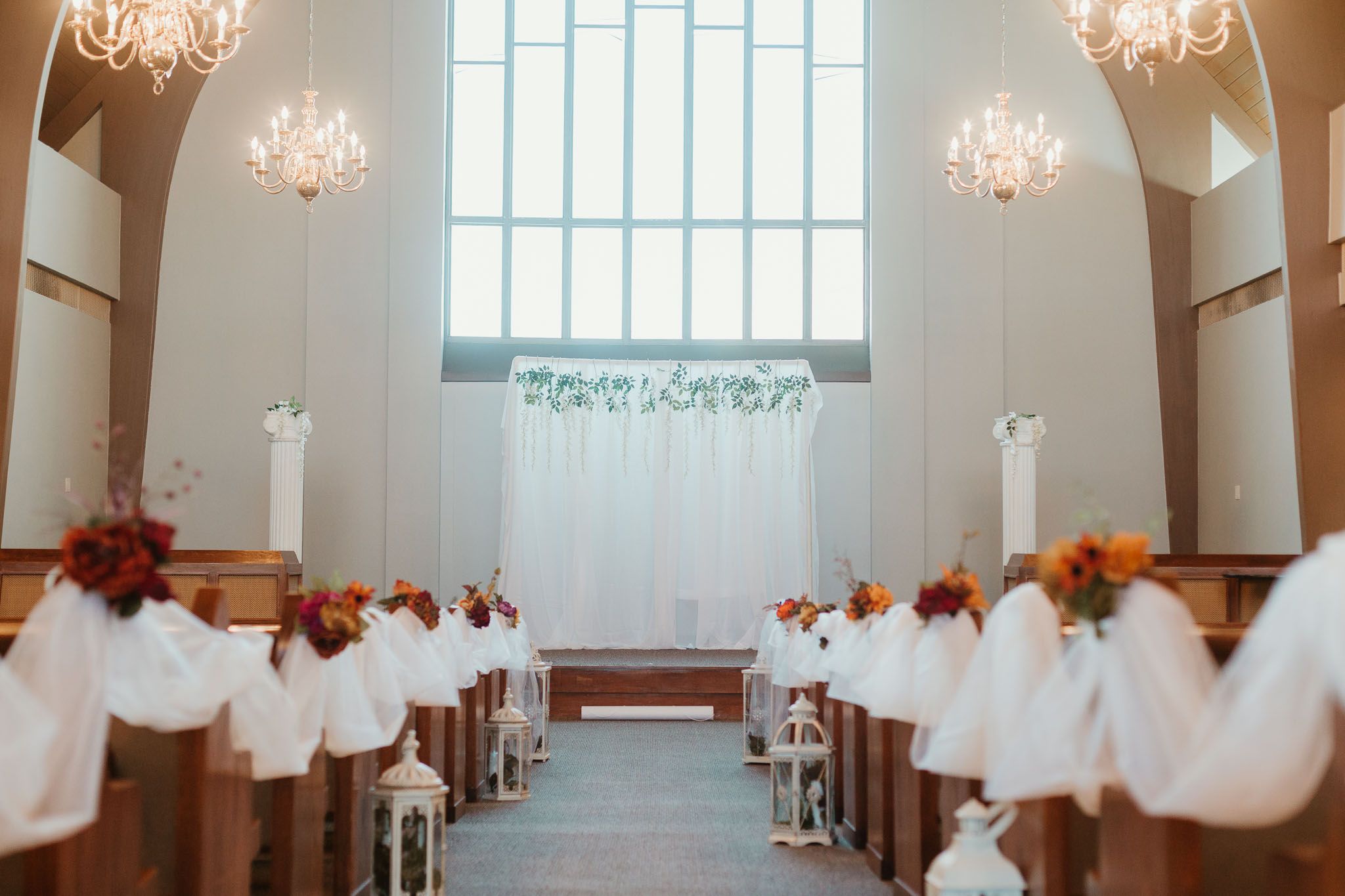 Elegant church wedding aisle decorated with white tulle, orange floral arrangements on wooden pews, and chandeliers.