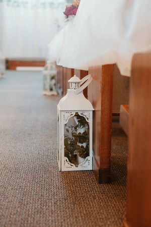 White decorative lantern with floral details placed on carpet beside wooden church pew at a wedding ceremony.