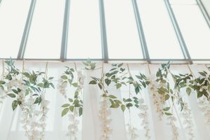 Elegant white floral garlands with green leaves hanging from a bright greenhouse ceiling for wedding decor.