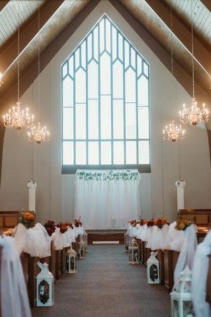 Elegant wedding chapel aisle with white pew decorations, lanterns, floral accents, chandeliers, and a large arched stained glass window.