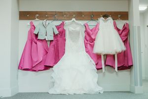 Wedding dress display with white bridal gown, pink bridesmaids dresses, flower girl dress, and gray suit jacket on hangers.