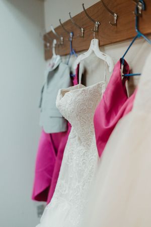 Wedding dress and pink bridesmaid gowns hanging on wooden hooks with a small gray suit in the background.