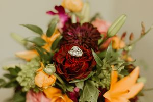Diamond engagement ring nestled in a dark red dahlia surrounded by vibrant orange and burgundy wedding bouquet flowers.