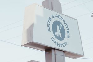 A white illuminated sign for an Arts and Activities Center with a bold K logo mounted on a pole outdoors.