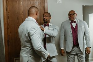Three groomsmen in light gray suits with burgundy bow ties helping each other prepare before a wedding ceremony.
