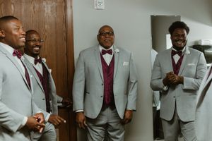 Groomsmen in gray suits with burgundy bow ties and vests laughing together before a wedding ceremony.