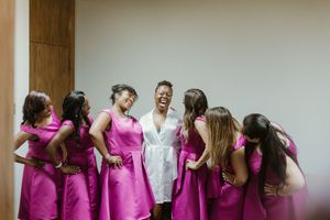 Bride in white robe laughing joyfully with bridesmaids in fuchsia pink dresses during wedding preparations.