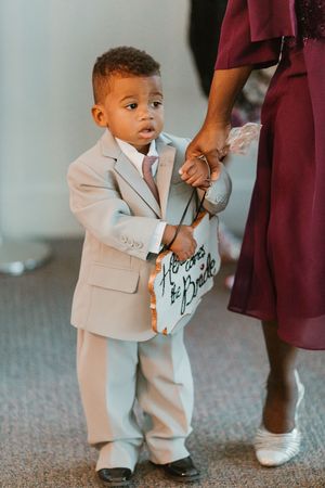 Adorable young ring bearer in a tan suit holds a sign reading Here Comes the Bride at a wedding ceremony.
