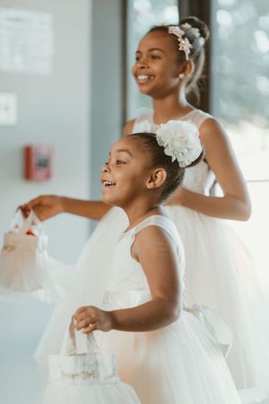 Two young Black flower girls in white tulle dresses smile and hold flower baskets at a wedding ceremony.