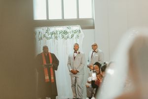 Groom stands at altar in gray suit during wedding ceremony with officiant and guests in bright venue.