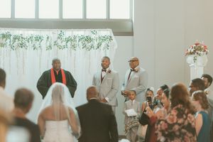 Groom and groomsmen in gray suits stand at altar during wedding ceremony with floral backdrop.
