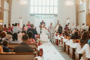 Wedding ceremony in a white chapel with bride and groom at the altar, guests seated in pews adorned with white ribbon and red flowers.
