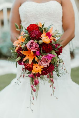 Bride holds a lush cascading wedding bouquet with red, orange, pink roses, lilies, and greenery.